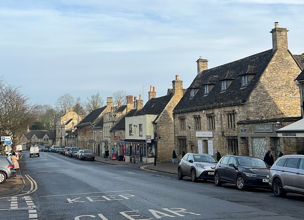 A street view of a charming village in the Cotswolds, featuring stone cottages and parked cars along the road.